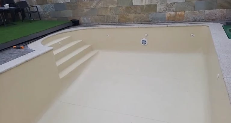 Empty inground swimming pool with cream-colored walls, built-in steps on the left side, and surrounding stone patio and landscaping